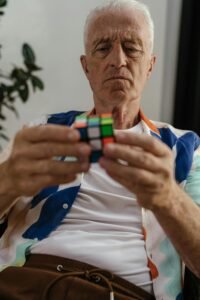 Elderly man focusing on solving a Rubik's Cube indoors, demonstrating concentration and puzzle-solving skills.