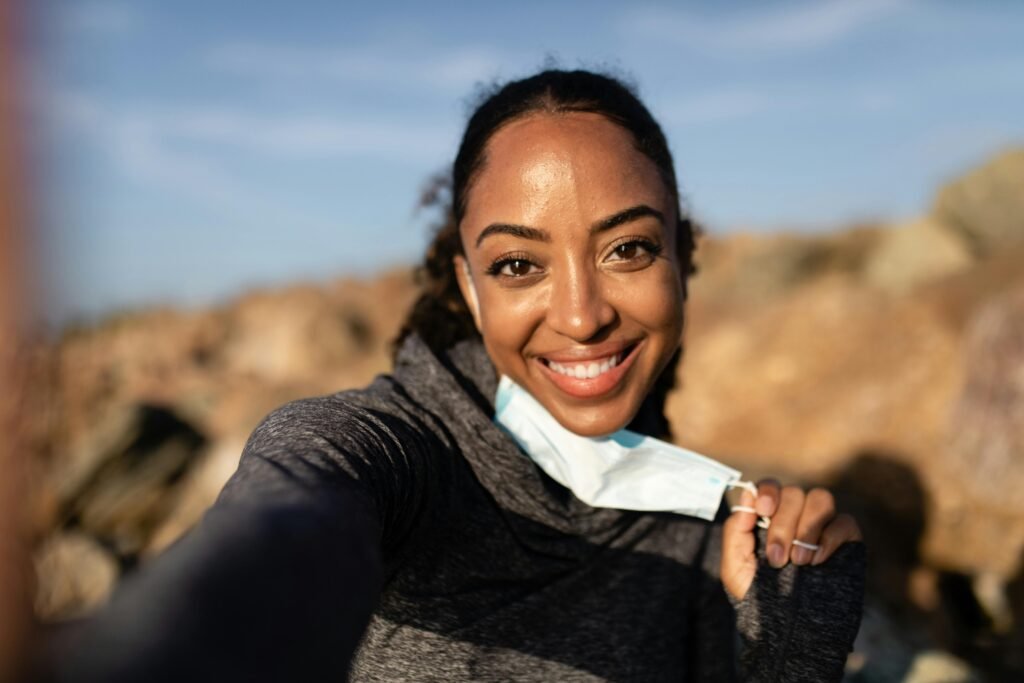 A cheerful woman taking a selfie outside, holding a face mask with a bright smile.