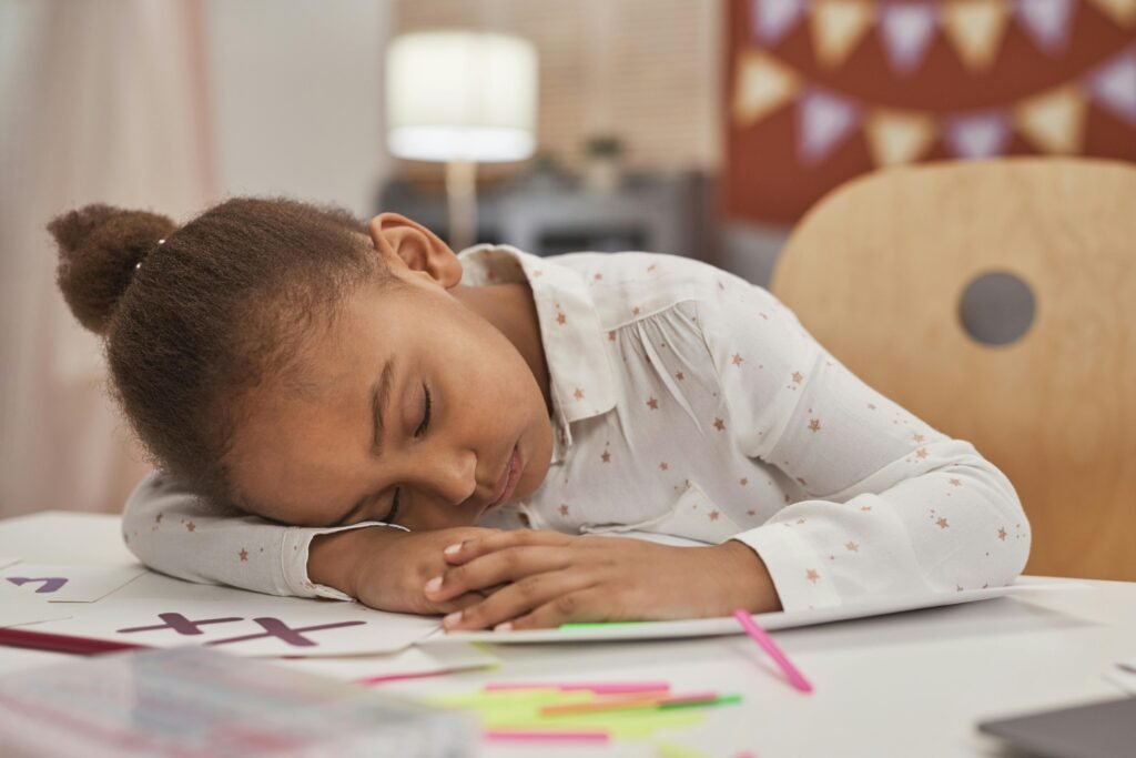 A tired young girl with closed eyes rests her head on homework at a table indoors.